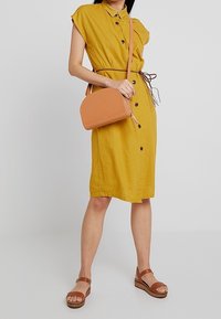 Šaty barvy hořčice s krátkými rukávy a zapínáním na knoflíky, s úzkým pasem, doplněné světlehnědou taškou přes rameno a hnědými sandály na pásku.