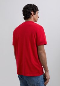 Man met kort krullend haar, draagt een effen rode t-shirt met korte mouwen en blauwe jeans, staat met de rug naar de kijker tegen een effen lichte achtergrond.