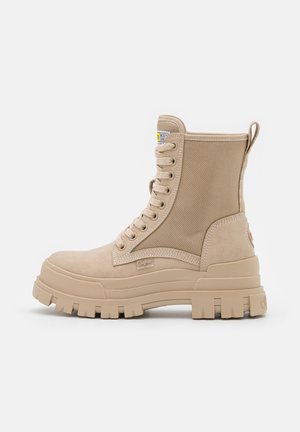 Bottine beige à lacets avec semelle épaisse à rainures et panneau latéral texturé, montrant l’étiquette de la marque et la languette à l’arrière.