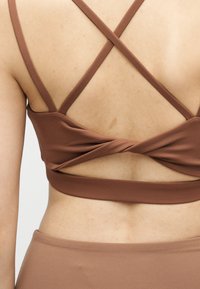 Οπίσθια όψη ατόμου που φοράει καφέ αθλητικό σουτιέν με λουριά σε σχήμα σταυρού και αντίστοιχα ψηλοκάβαλα σορτς.