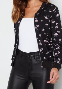 Femme portant une veste zippée noire à fleurs, un débardeur blanc à côtes et un pantalon en simili cuir noir, main visible avec des ongles rose clair.