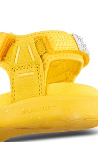 Keltainen sandaali, jossa on säädettävät kangasnauhat, pinta-aktiiviset mokka-aksentit ja kumipohja, johon on painettu "NATURAL SOFT™."
