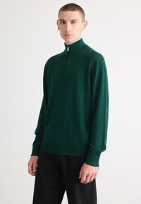 Maglione a girocollo verde scuro con colletto alto e zip frontale. Realizzato in morbido tessuto a maglia, presenta maniche lunghe e una silhouette aderente.