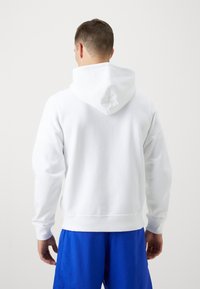 Hvit hettegenser med snor foran, ribbede mansjetter og nederkant. Båret med blå treningsshorts, som viser en myk stoffstruktur.