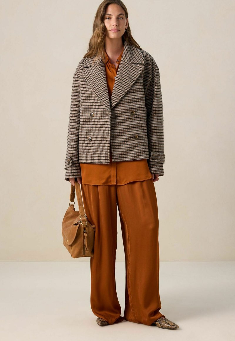 Femme portant un manteau à carreaux marron à double boutonnage sur un pantalon ample rouille et une chemise, tenant un sac à main beige, se tenant sur un fond neutre.