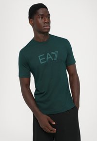 Jauns vīrietis valkā tumši zaļu Emporio Armani EA7 T-kreklu un melnas bikses, stāvot pretī vienkāršai baltai fona.