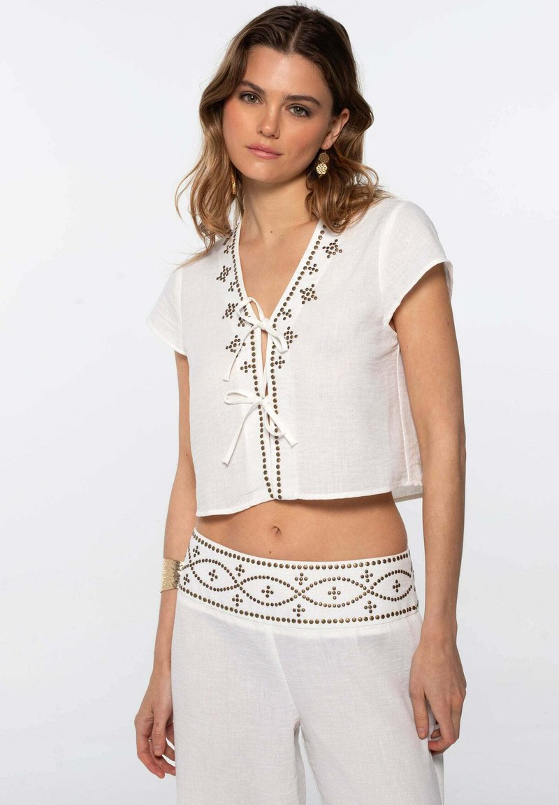 Top corto de lino blanco con mangas cortas, escote en forma de V y decoración de cuentas a lo largo del escote y el dobladillo. Combinado con pantalones blancos sueltos a juego.