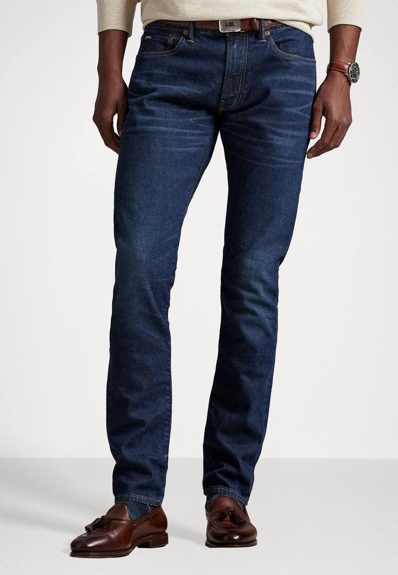 Dark blue denim jeans with a slim fit, subtle fading, and stitched accents. Paired with brown leather loafers and a wristwatch.