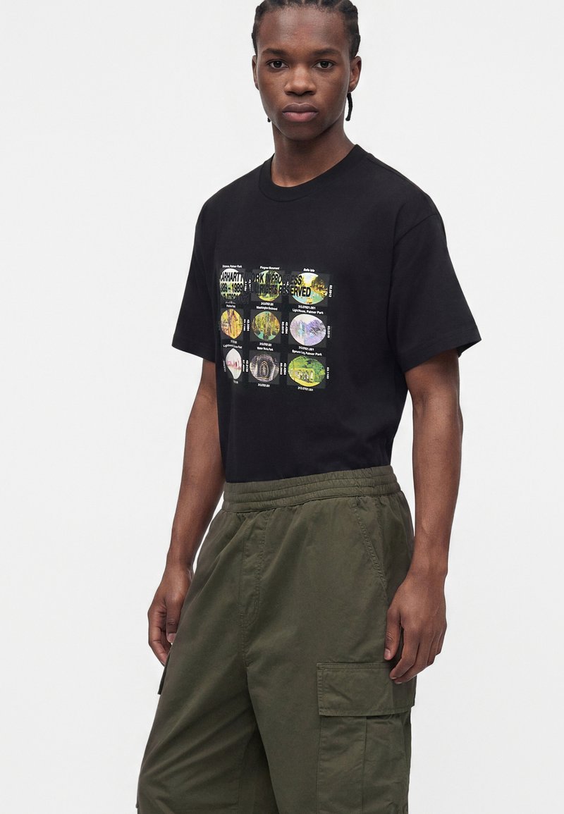 Junger Mann trägt ein schwarzes T-Shirt mit bunten kreisförmigen Motiven und olivgrünen Cargohosen, steht vor einem schlichten Hintergrund.