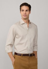 Camisa de botones de color claro con un patrón a cuadros, mangas largas arremangadas hasta los codos, combinada con pantalones color beige y un cinturón marrón.