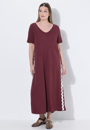 Kvinde iført en vinrød maxikjole med korte ærmer, hvide bølgede striber på siderne og brune sandaler, stående foran en helt hvid baggrund.