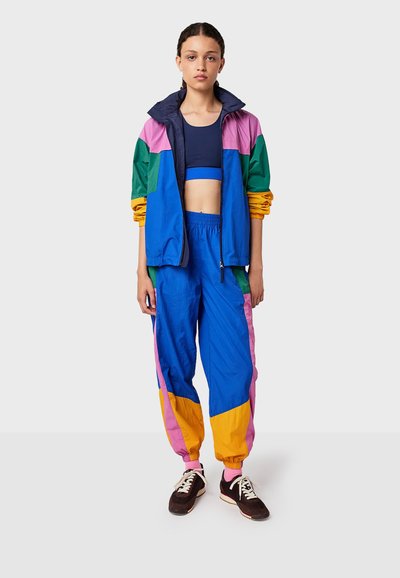 Kleurrijke trainingsoutfit met een blauwe en groene jas met roze en gele accenten, gecombineerd met comfortabele blauwe broeken en zwarte sneakers.