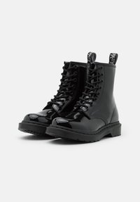 Botas de tobillo negras brillantes con cordones, suelas gruesas y lengüetas de marca sobre un fondo blanco.