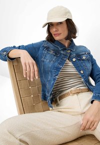 Giacca di denim blu con tasche frontali, top beige a righe, pantaloni crema strutturati e cappellino da baseball chiaro. Modella seduta su una sedia di legno.