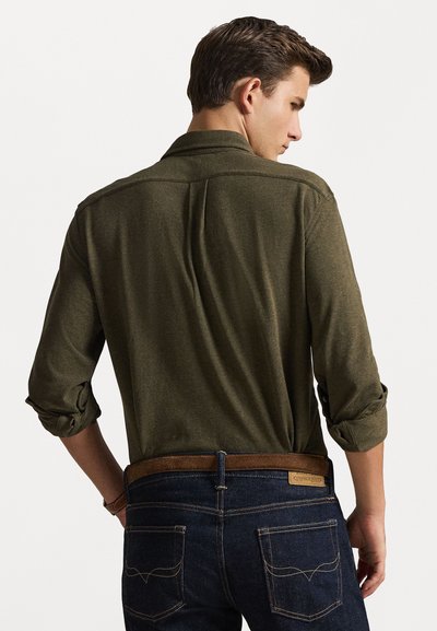 Camicia verde oliva a maniche lunghe con maniche arrotolate, abbinata a jeans blu scuro con una cintura in pelle marrone e tasche posteriori cucite.