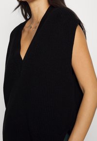 Schwarzes, gestricktes, ärmelloses Oberteil mit tiefem V-Ausschnitt, strukturiertem Stoff und seitlichen Schlitzen für Atmungsaktivität. Goldene Halskette als Accessoire.