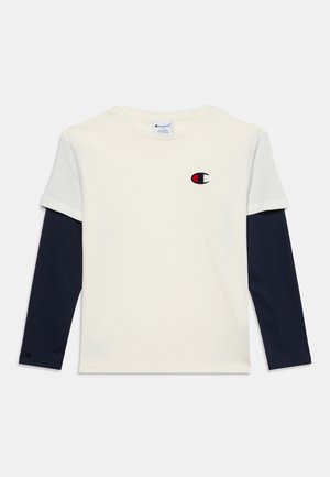 T-shirt Champion crème à manches courtes porté sur un haut bleu marine à manches longues, avec un petit logo sur la poitrine.