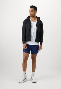 Pánský atletický outfit sestávající z černé zipové bundy, bílé tričko, modrých šortek a bílých tenisek s černými akcenty.