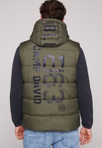 Gilet imbottito verde oliva con stampa blu notte che presenta la scritta "CAMP DAVID" e le coordinate. Progettato con cappuccio e tessuto texturizzato, abbinato a un maglione blu scuro.