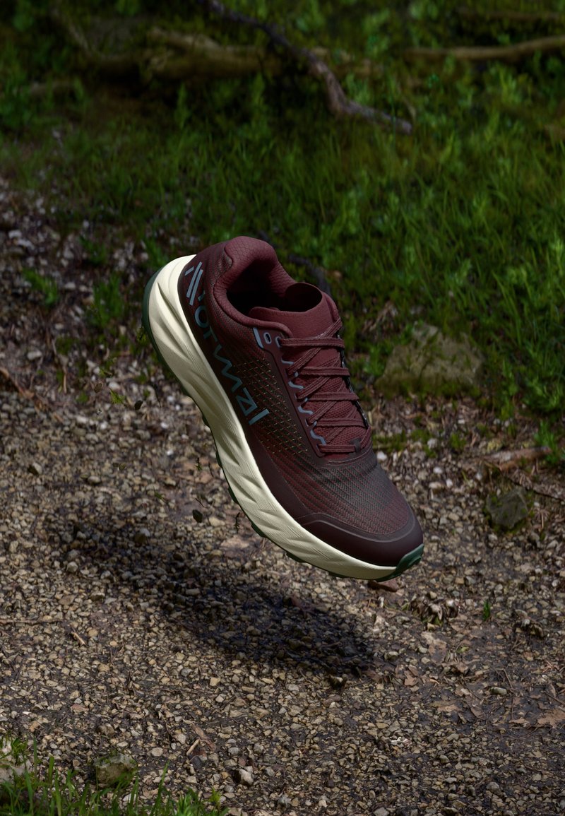 Maroon running shoe with a textured upper, laces, and a cushioned cream-colored sole, shown floating above a gravel path amidst greenery.