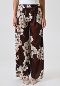 Brede, bruine bloemenprintbroeken van gladde stof met roomwitte en roze bloemen; heeft plooien en een vloeiende uitstraling.