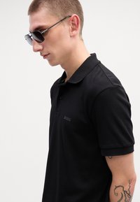 Melns polo krekls ar smalku logo uz krūtīm, klasiskā apkaklē un īsām piedurknēm. Komplektā ar tumšiem saulesbrillēm.