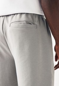 Gros plan sur la hanche d'une personne montrant un pantalon gris clair avec une poche arrière et un petit logo cousu près de la taille.