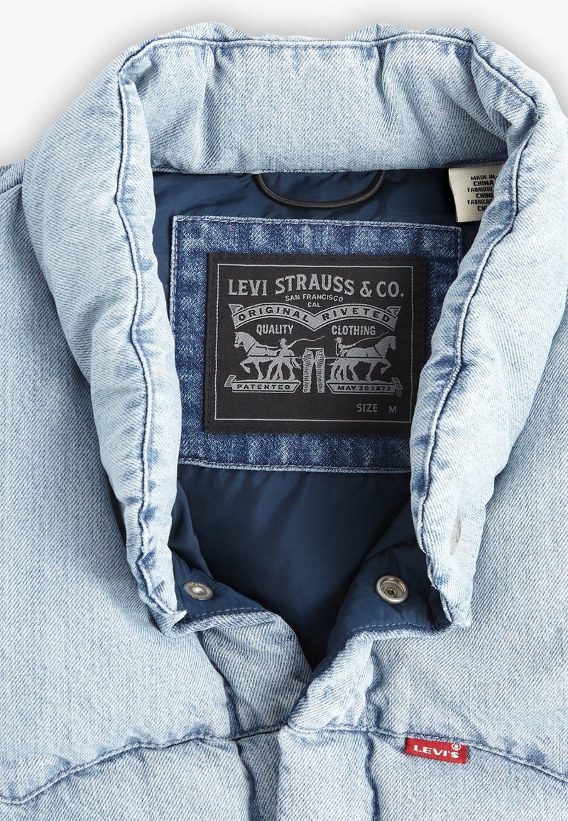 Veste en denim bleu clair avec un col, ornée d'une étiquette noire avec un design et d'un petit tag rouge Levi's à l'avant. Texture lisse.