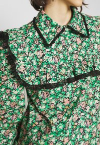 Femme portant un chemisier à fleurs avec des motifs verts, roses et blancs, orné de dentelle noire et de boutons en perles sur un col pointu.