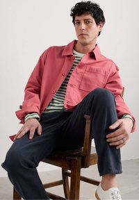 Veste rouge rubis avec une poche, portée sur une chemise rayée verte et blanche, assortie à un pantalon noir et des chaussures blanches, assise sur une chaise en bois.