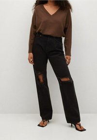 Brązowy sweter z dekoltem w serek w połączeniu z czarnymi podartymi dżinsami. Spodnie mają przetarcia na kolanach oraz prosty krój nogawek i są uzupełnione czarnymi sandałami.