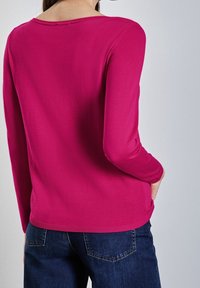 Fuksia pitkähihainen paita pyöreällä pääntiellä, pehmeällä tekstuurilla ja resorilla helmassa, yhdistetty tummansinisiin farkkuihin.