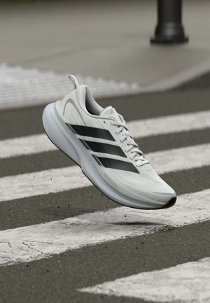 Scarpa sportiva bianca con strisce nere, design leggero, superficie liscia, dotata di suola imbottita e un anello per indossarla facilmente, fotografata a mezz'aria.