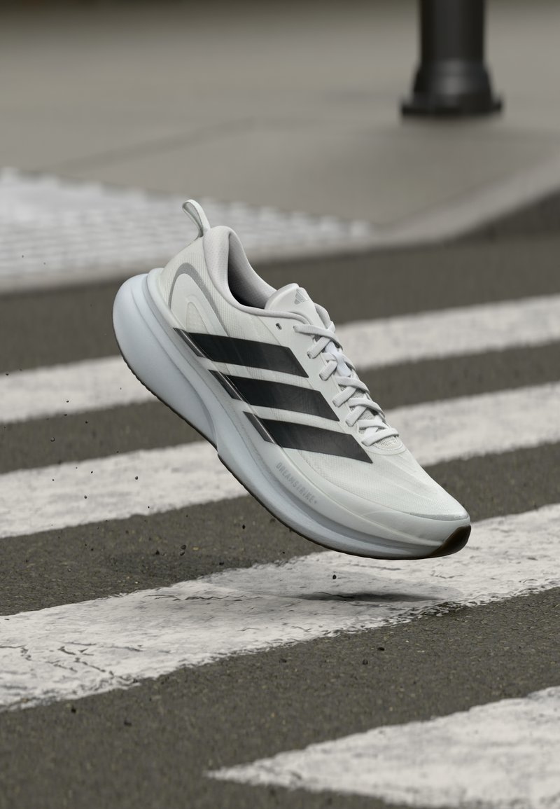 Scarpa sportiva bianca con strisce nere, design leggero, superficie liscia, dotata di suola imbottita e un anello per indossarla facilmente, fotografata a mezz'aria.