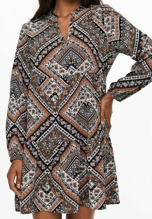Femme portant une robe à manches longues avec un motif géométrique et floral en noir, blanc, orange et beige.