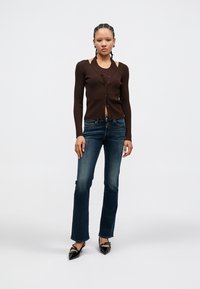 Rjav ribičast kardigan s sponko na gumbe, kombiniran s temno modrimi razširjenimi kavbojkami in črnim čevljem s špičastim nožnim delom. Model stoji na svetlem ozadju.