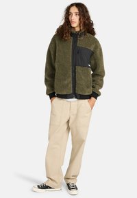Veste en polaire vert olive avec des accents en nylon noir, fermeture zippée et poignets côtelés, associée à un pantalon beige décontracté et des baskets noires.