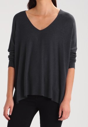 Mujer con un suéter de manga larga y cuello en V holgado de color gris oscuro combinado con pantalones ajustados negros, posando frente a un fondo liso.