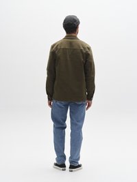 Cămașă verde măsliniu cu nasturi, croială dreaptă, asortată cu blugi din denim albastru deschis și adidași negri, văzută din spate.