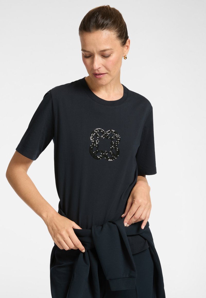 Femme portant un t-shirt noir avec un motif de fleurs en sequins argentés et noirs, nouant un vêtement noir assorti autour de sa taille.