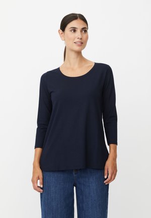 Frau mit dunklem, zurückgebundenem Haar, die ein marineblaues Langarmshirt und blaue Jeans trägt, steht vor einem schlichten weißen Hintergrund.