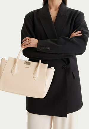 Donna con blazer nero che tiene una borsa rettangolare color crema con manici superiori e chiusura metallica.
