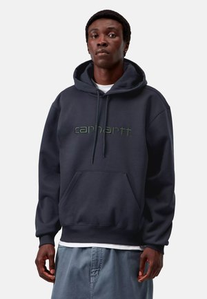 Man wearing a dark blue Carhartt hoodie with front pocket and drawstrings, standing against a plain light background.