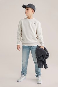 Krémově texturovaná mikina s geometrickým vzorem, světle modré džínové kalhoty a šedá čepice, držící tmavě šedou bundu. Bílá teniska doplňuje vzhled.