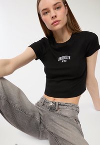 Crop top nero a coste con testo "BROOKLYN NYC" in bianco, abbinato a jeans grigi a vita alta con una texture scolorita.