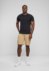 T-shirt noir à manches courtes associé à un short cargo kaki doté de plusieurs poches, porté avec des baskets blanches et des chaussettes de mi-mollet visibles.
