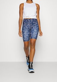 Top bianco senza maniche con un nodo sul davanti, abbinato a pantaloni bici blu navy con motivi a righe bianche. Il modello indossa sneaker alte nere e blu.