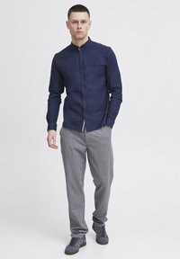 Camisa azul marino de botones con cuello alto, combinada con pantalones de chándal gris claro y zapatillas grises. La camisa cuenta con un bolsillo en el pecho y un corte ajustado.