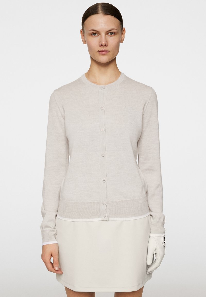Ljusgrå cardigan, stickad textur, rund halsringning, sju framknappar, figurnära design, ribbade mudd och nederkant, minimalistisk logotyp på bröstet.