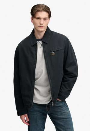 Ung mand iført en sort lynlåstrøje over en lysegrå skjorte og blå jeans, står med den ene hånd i lommen.